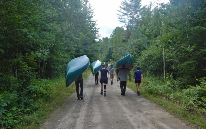 Three upside-down canoes are carried by students along a trail in a wooded area